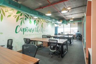 An open office workspace featuring several desks with ergonomic chairs. The walls are decorated with a nature-themed mural and the phrase 'Capture the Nature' is prominently displayed. Overhead, modern light fixtures and exposed ductwork add an industrial touch. Natural light filters in through large windows, enhancing the workspace.
