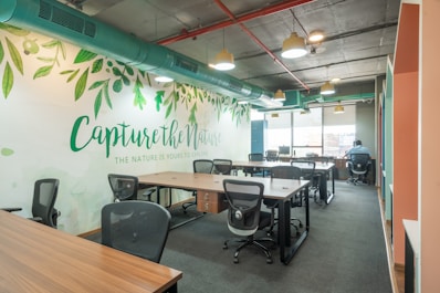 An open office workspace featuring several desks with ergonomic chairs. The walls are decorated with a nature-themed mural and the phrase 'Capture the Nature' is prominently displayed. Overhead, modern light fixtures and exposed ductwork add an industrial touch. Natural light filters in through large windows, enhancing the workspace.