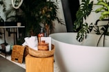 A modern and stylish bathroom setup featuring a white oval bathtub surrounded by lush green plants. In the foreground, a wicker basket on a wooden stool contains various bath items such as bottles of soap, a brush, and white towels. The background includes a dark table with a potted plant, wicker baskets, and more greenery, creating a natural and serene atmosphere.