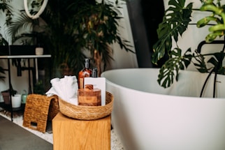 A modern and stylish bathroom setup featuring a white oval bathtub surrounded by lush green plants. In the foreground, a wicker basket on a wooden stool contains various bath items such as bottles of soap, a brush, and white towels. The background includes a dark table with a potted plant, wicker baskets, and more greenery, creating a natural and serene atmosphere.