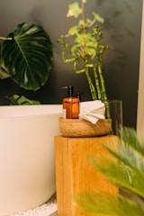A serene bathroom setting with a white bathtub partially visible. A wooden stool holds a woven basket containing a brown bottle with a pump dispenser and folded white towels. Large, vibrant green leaves and bamboo stalks add a touch of nature to the scene. White pebbles are scattered below, enhancing the spa-like atmosphere.