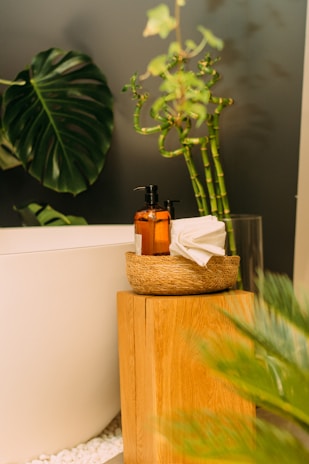 A serene bathroom setting with a white bathtub partially visible. A wooden stool holds a woven basket containing a brown bottle with a pump dispenser and folded white towels. Large, vibrant green leaves and bamboo stalks add a touch of nature to the scene. White pebbles are scattered below, enhancing the spa-like atmosphere.