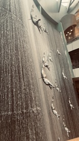 A towering indoor waterfall features several metallic sculptures of human figures diving in a vertical formation. The water cascades down a textured wall, creating a dramatic backdrop for the art installation. The setting appears to be inside a large modern building with a high ceiling and geometric architecture.