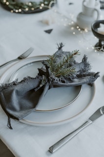 A table setting features a stack of neatly arranged plates with a dark fringed napkin on top, decorated with a sprig of evergreen. Silver cutlery is positioned on either side of the plates. The scene is softly lit with string lights, contributing to an elegant and cozy atmosphere.