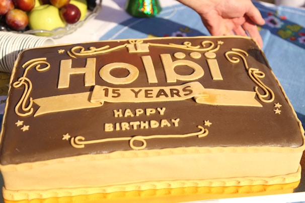 A rectangular cake with decorative elements and embossed text celebrating 15 years, surrounded by fruits and plates. A hand is positioned near the top of the cake.