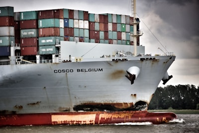 A large cargo ship with the name 'COSCO Belgium' on its hull is navigating through the water. The vessel is heavily loaded with stacked colorful shipping containers that are labeled with various brands. The ship shows signs of wear, with rust visible near the waterline. The background features a cloudy sky and a strip of land with trees.