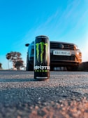A can of Monster Energy drink is prominently placed on a textured road surface. Behind it, a car is positioned slightly out of focus, with a license plate visible. The scene is set outdoors with a bright blue sky and a soft glow from the sun, likely during sunset or sunrise.