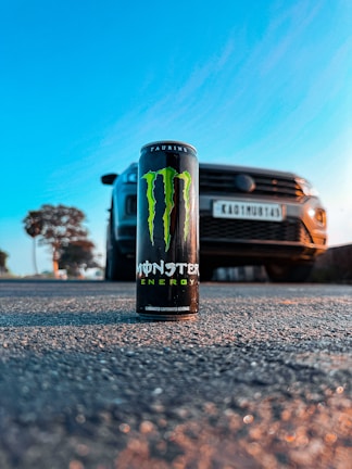 A can of Monster Energy drink is prominently placed on a textured road surface. Behind it, a car is positioned slightly out of focus, with a license plate visible. The scene is set outdoors with a bright blue sky and a soft glow from the sun, likely during sunset or sunrise.