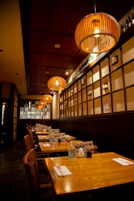 A cozy restaurant interior with wooden tables and chairs lined up along the wall. Warm, round pendant lights hang from the ceiling, creating a welcoming ambiance. The wall features a grid pattern with subtle decorations and bottles on the top shelf. Each table is neatly set with menus, napkins, and small condiment bottles.