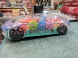A transparent toy car revealing colorful interlocking gears inside. The vehicle is placed on a textured surface, with various other objects and boxes visible in the background.