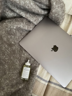 A cozy and aesthetic composition featuring a silver laptop partially covered by a soft, textured gray blanket. Next to the laptop, there is a bottle of perfume labeled 'Replica' placed on the blanket. The background includes a beige and black striped fabric, adding to the warm and inviting atmosphere.