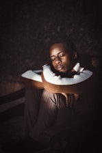 A person is sitting with eyes closed, resting their chin on their crossed arms. They are wearing a striped sweater and pinstripe pants, seated on a chair in a dimly lit setting.