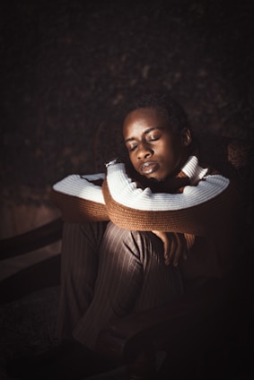 A person is sitting with eyes closed, resting their chin on their crossed arms. They are wearing a striped sweater and pinstripe pants, seated on a chair in a dimly lit setting.