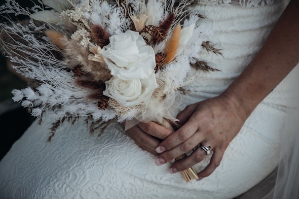 A bouquet composed of white roses, dried flowers, and various types of grasses is held by a person in a white lace dress. The hands, adorned with rings, gently cradle the bouquet. The textures of the lace and dried flowers create a rustic and elegant feel.