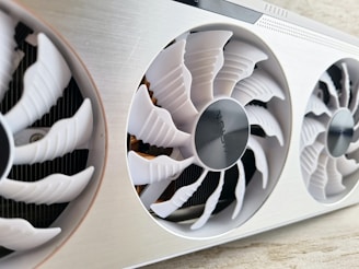 A close-up view of a computer graphics card with three large, white cooling fans enclosed in a metallic casing. The central fan has a visible logo and intricate blade design.