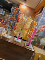 Three colorful candy stands are displayed on a wooden counter. Each stand holds a variety of lollipops, including spirals, swirls, and heart shapes in vibrant colors like red, blue, green, yellow, and orange. The background features a softly lit interior with shelves displaying more confectionery items.