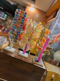 Three colorful candy stands are displayed on a wooden counter. Each stand holds a variety of lollipops, including spirals, swirls, and heart shapes in vibrant colors like red, blue, green, yellow, and orange. The background features a softly lit interior with shelves displaying more confectionery items.