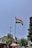 A national flag is prominently displayed on a tall flagpole against a clear blue sky. It is situated in an outdoor environment with trees, a streetlamp, and various structures including a small building and market stalls covered with colorful umbrellas.