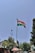 A national flag is prominently displayed on a tall flagpole against a clear blue sky. It is situated in an outdoor environment with trees, a streetlamp, and various structures including a small building and market stalls covered with colorful umbrellas.