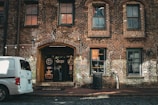 A rustic brick building with arched windows and a prominent entrance labeled with signage for a coffee company. A white van is parked on the cobblestone street in front. The setting is urban with an old-fashioned charm, featuring visible light fixtures and a trash can on the sidewalk.