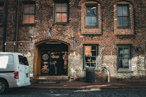 A rustic brick building with arched windows and a prominent entrance labeled with signage for a coffee company. A white van is parked on the cobblestone street in front. The setting is urban with an old-fashioned charm, featuring visible light fixtures and a trash can on the sidewalk.
