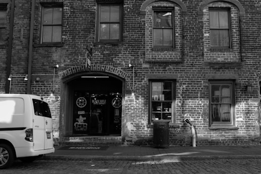 A brick building facade with arched windows and a door with signage above it, including 'BLACK RIFLE COFFEE COMPANY.' A white van is parked on the cobblestone street in front, and there's a trash can nearby.