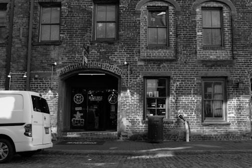 A brick building facade with arched windows and a door with signage above it, including 'BLACK RIFLE COFFEE COMPANY.' A white van is parked on the cobblestone street in front, and there's a trash can nearby.