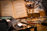 A vintage setting featuring an open book with text in a foreign language. Several magnifying glasses with different handle designs are spread out on a wooden surface. In the background, ornate pens are arranged in a white holder, creating a cozy, scholarly atmosphere.