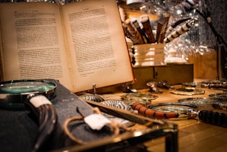 A vintage setting featuring an open book with text in a foreign language. Several magnifying glasses with different handle designs are spread out on a wooden surface. In the background, ornate pens are arranged in a white holder, creating a cozy, scholarly atmosphere.