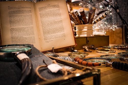 A vintage setting featuring an open book with text in a foreign language. Several magnifying glasses with different handle designs are spread out on a wooden surface. In the background, ornate pens are arranged in a white holder, creating a cozy, scholarly atmosphere.