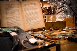A vintage setting featuring an open book with text in a foreign language. Several magnifying glasses with different handle designs are spread out on a wooden surface. In the background, ornate pens are arranged in a white holder, creating a cozy, scholarly atmosphere.