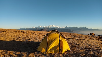 A yellow tent is set up on a grassy plateau with a backdrop of majestic snow-capped mountains under a clear blue sky. In the distance, a few horses are grazing, adding to the serene and natural setting.