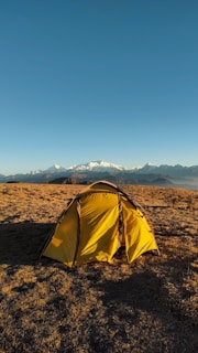 A yellow tent is set up on a grassy field under a clear blue sky, with snowy mountain peaks visible in the distance. The scene suggests an outdoor camping experience in a serene, natural environment.