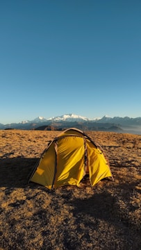 A yellow tent is set up on a grassy field under a clear blue sky, with snowy mountain peaks visible in the distance. The scene suggests an outdoor camping experience in a serene, natural environment.