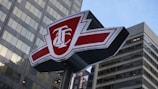 A red and white sign with the initials T.B. standing prominently in front of tall, modern skyscrapers. The sign has a bold design with sharp angles and is perched atop a metal pole.