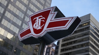 A red and white sign with the initials T.B. standing prominently in front of tall, modern skyscrapers. The sign has a bold design with sharp angles and is perched atop a metal pole.