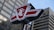 A red and white sign with the initials T.B. standing prominently in front of tall, modern skyscrapers. The sign has a bold design with sharp angles and is perched atop a metal pole.