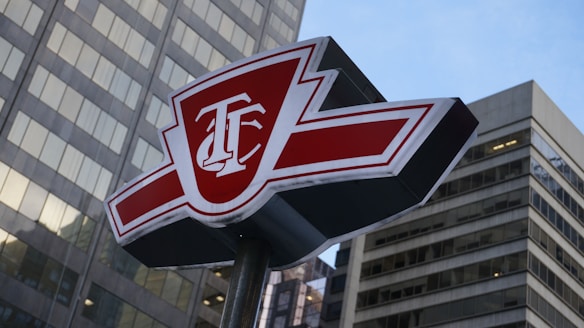 A red and white sign with the initials T.B. standing prominently in front of tall, modern skyscrapers. The sign has a bold design with sharp angles and is perched atop a metal pole.