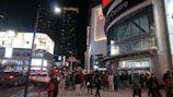 A bustling urban street scene at night with pedestrians walking by a shopping center. The streets are brightly lit by billboard advertisements, and the entrance to the Eaton Centre is visible. Several people are dressed warmly, indicating cold weather. There are vehicles on the street and high-rise buildings in the background.