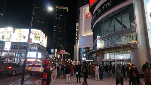 A bustling urban street scene at night with pedestrians walking by a shopping center. The streets are brightly lit by billboard advertisements, and the entrance to the Eaton Centre is visible. Several people are dressed warmly, indicating cold weather. There are vehicles on the street and high-rise buildings in the background.