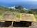 Two wooden plaques with inscriptions in English and Maori stand on a grassy cliffside overlooking a vast ocean and rolling hills. The plaques appear weathered, suggesting exposure to the coastal elements. The scenic backdrop of the blue sea fades into the horizon under a clear sky, with rugged greenery covering the landscape.