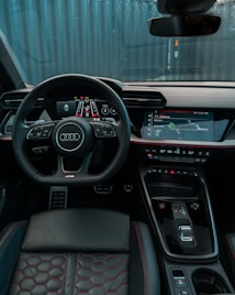 A modern car interior featuring a sleek, black dashboard with digital displays and controls. There's a prominent steering wheel with an Audi logo, and the seats are upholstered in black leather with red stitching. The overall design is high-tech and ergonomic.