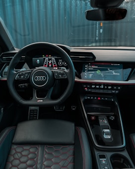 A modern car interior featuring a sleek, black dashboard with digital displays and controls. There's a prominent steering wheel with an Audi logo, and the seats are upholstered in black leather with red stitching. The overall design is high-tech and ergonomic.