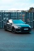 A sleek, modern car with a dark gray finish is parked in front of large industrial containers. The car's design features sharp lines and sporty elements, including black wheels and a detailed front grille. The background consists of metal containers with visible text and numbers, suggesting a storage or shipping environment.