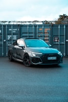 A sleek, modern car with a dark gray finish is parked in front of large industrial containers. The car's design features sharp lines and sporty elements, including black wheels and a detailed front grille. The background consists of metal containers with visible text and numbers, suggesting a storage or shipping environment.