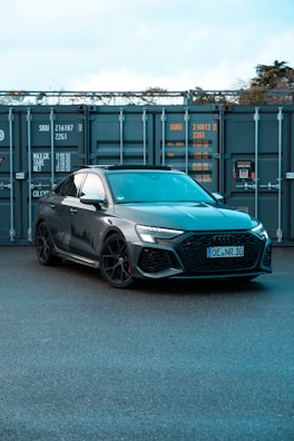 A sleek, modern car with a dark gray finish is parked in front of large industrial containers. The car's design features sharp lines and sporty elements, including black wheels and a detailed front grille. The background consists of metal containers with visible text and numbers, suggesting a storage or shipping environment.