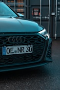 The image features the front end of a car with a visible license plate reading 'OE NR 30'. The car has a sleek black grille and is parked in front of what appears to be an industrial or shipping container backdrop.
