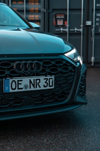 The image features the front end of a car with a visible license plate reading 'OE NR 30'. The car has a sleek black grille and is parked in front of what appears to be an industrial or shipping container backdrop.