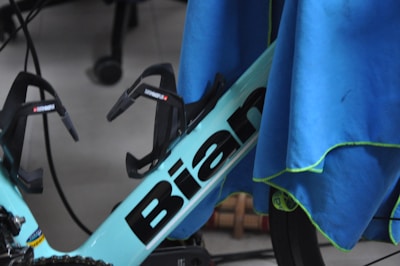 A section of a bicycle frame is partially covered by a blue cloth. The visible segment of the frame is labeled with the word 'Bian' and includes two black bottle holders attached. The surrounding area includes parts of a wheel and other cycling components, with some wires in the background.