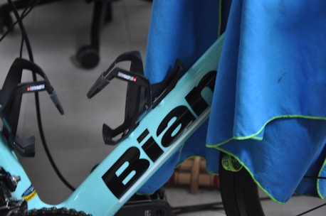 A section of a bicycle frame is partially covered by a blue cloth. The visible segment of the frame is labeled with the word 'Bian' and includes two black bottle holders attached. The surrounding area includes parts of a wheel and other cycling components, with some wires in the background.
